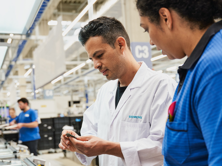 Ein Herr mit einer weißen Jacke und eine Frau in einem blauen T-Shirt sehen auf ein kleines Bauteil.